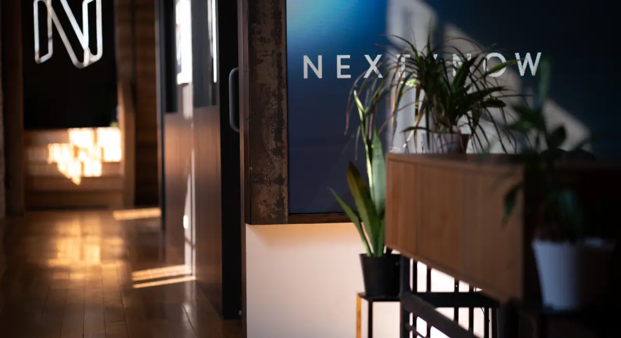 Picture of the Next/Now office, logos in background, plants in foreground